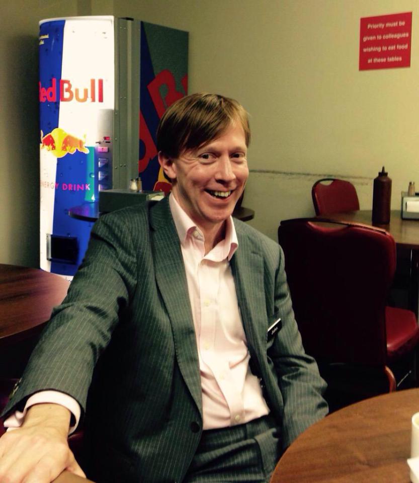 Man in suit sitting at a table near a Red Bull vending machine.