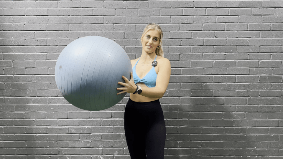 a woman in a blue top holds a pilates ball in front of a brick wall