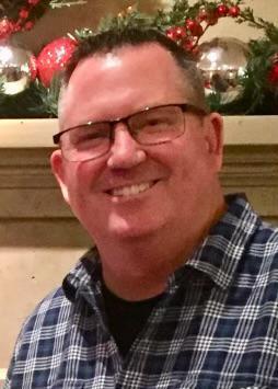 a man wearing glasses and a plaid shirt is smiling in front of a fireplace .
