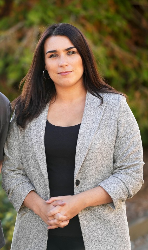 a woman in a grey jacket stands with her hands folded