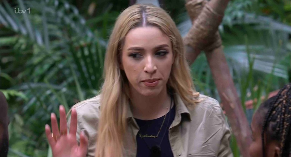 A blonde woman waves her hand in a jungle setting.