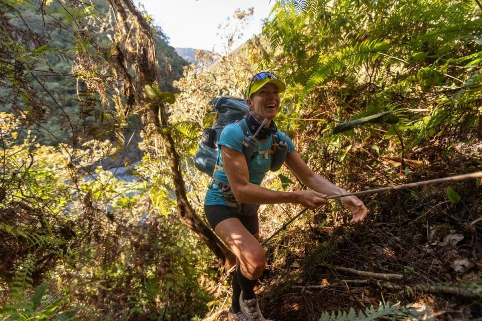A smiling woman hiking through lush forest, using a rope to assist her ascent.