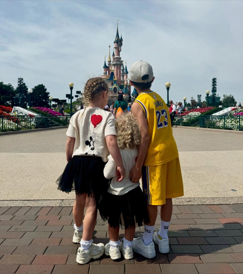 Three children stand facing a Disney castle.