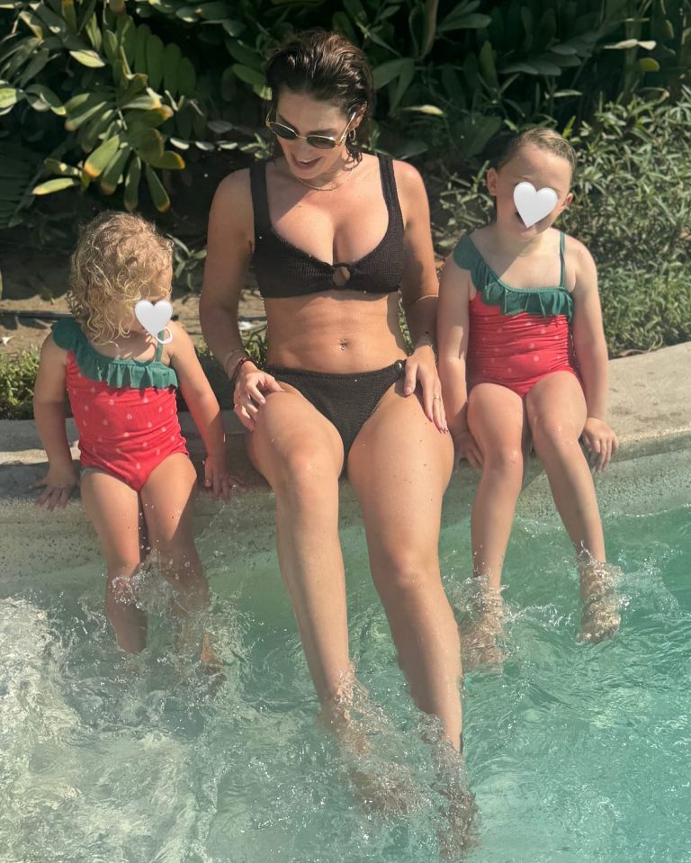 Woman in a black bikini with two young girls in red swimsuits at a pool.