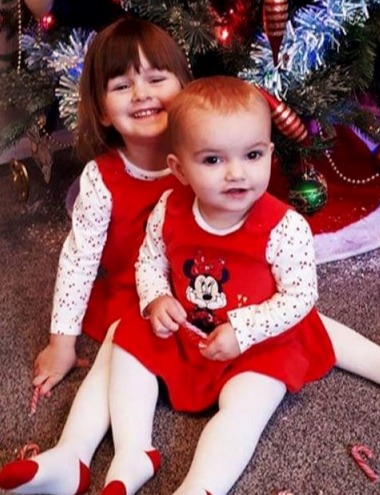 Two young girls in red Christmas dresses sitting in front of a decorated Christmas tree.