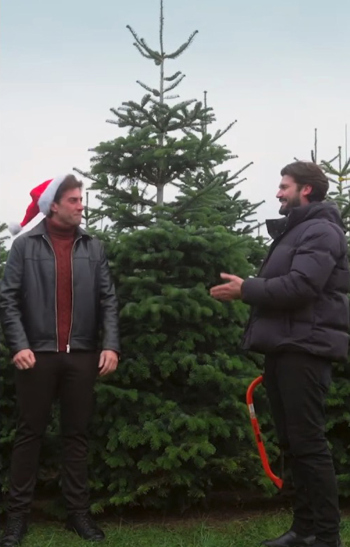Two men stand by Christmas trees; one wears a Santa hat.