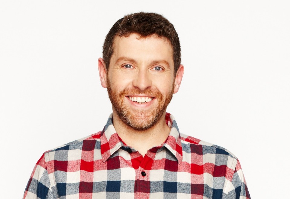 Dave Gorman in jeans and a red and blue plaid shirt.