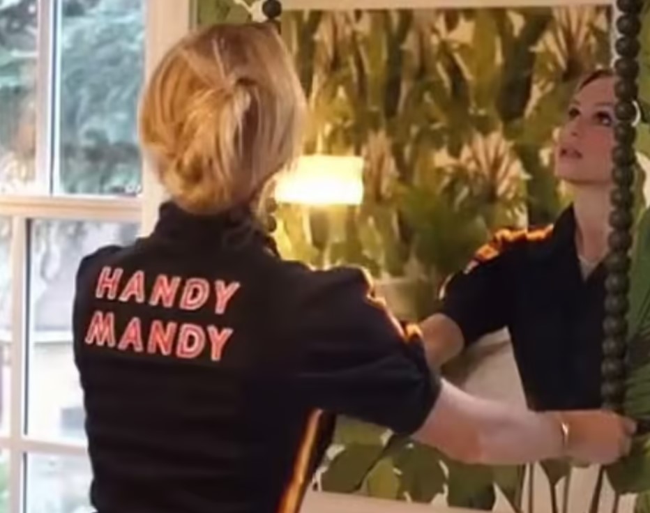 Amanda Holden wearing a "Handy Mandy" jumpsuit looks in a mirror.