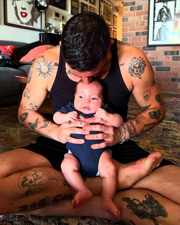 A father with many tattoos kisses his baby son on the forehead.