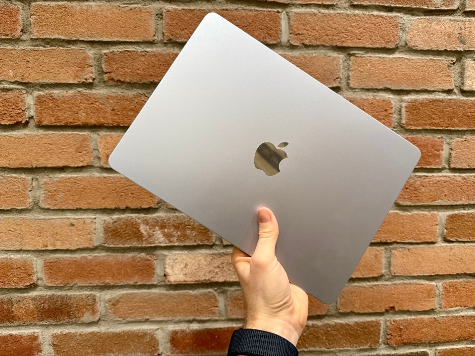 A hand holding a silver Apple laptop against a brick wall.