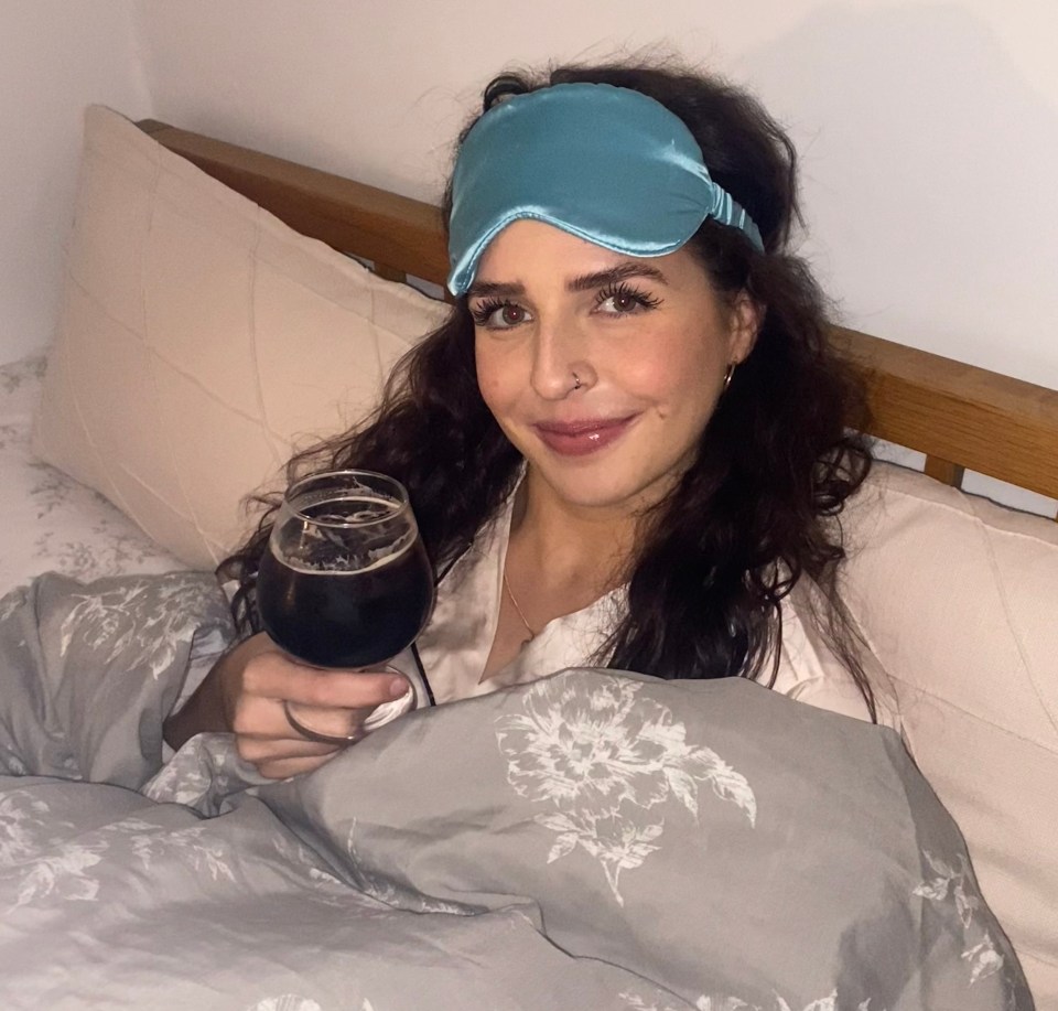 Woman in bed wearing a sleep mask, holding a glass of dark liquid.