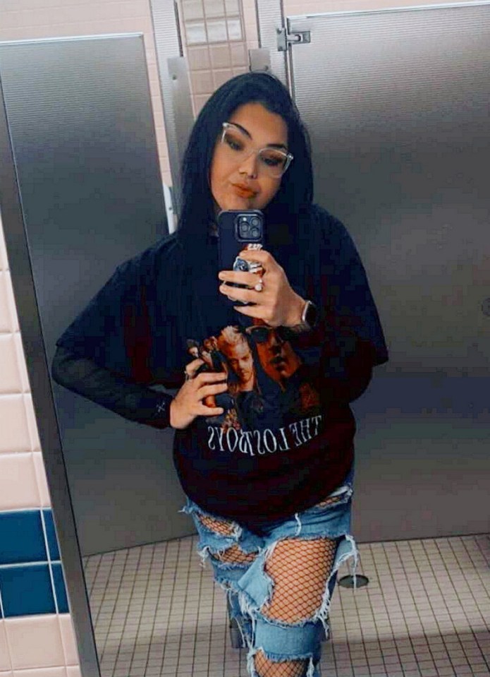 Woman taking a selfie in a restroom wearing a "The Lost Boys" t-shirt and ripped jeans.