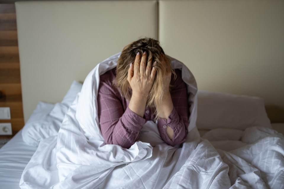 Woman sitting in bed, head in hands, appearing distressed.