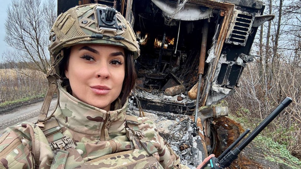 A female soldier in camouflage uniform stands near a destroyed military vehicle.