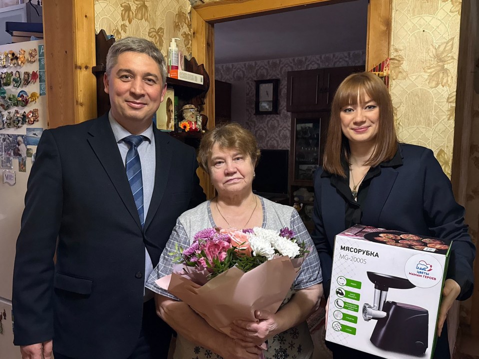 A woman receives a meat grinder and flowers from two people.