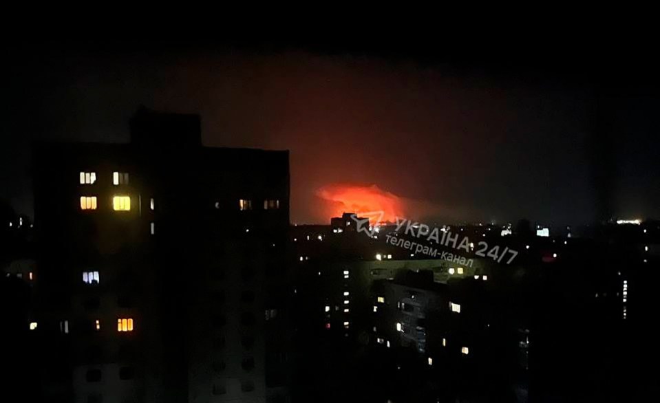 Nighttime view of a city with a large fire or explosion in the distance.