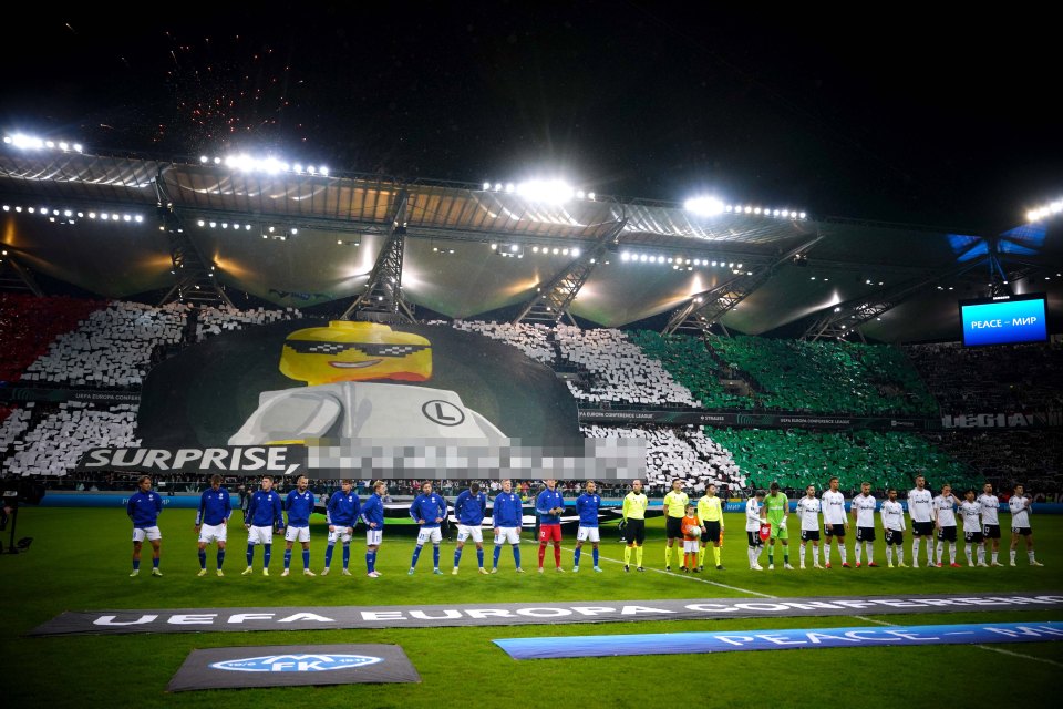 Legia Warsaw and Molde soccer teams line up before a match, with a large Lego-themed banner displayed in the stands.