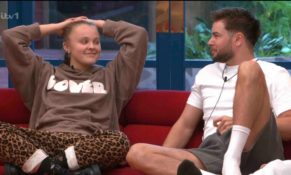Two people sitting on a red couch; a woman in a leopard print pants and a brown hoodie, and a man in a white shirt and gray shorts.