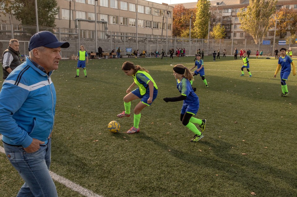 Girls' soccer team playing on a field, with their coach watching.