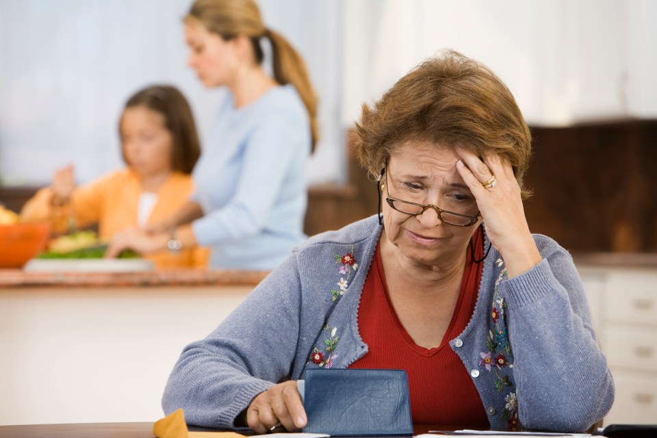 A worried grandmother looks at bills with her granddaughter and daughter in the background.