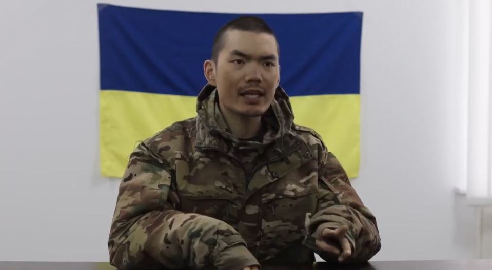 A Chinese soldier in Ukraine speaks in front of a Ukrainian flag.