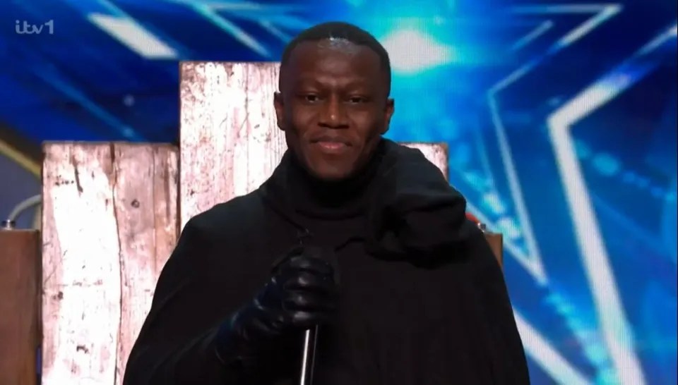 Screenshot of a man in a dark hooded cloak holding a microphone.