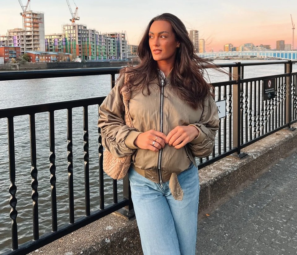 Woman in bomber jacket by river, buildings under construction in background.