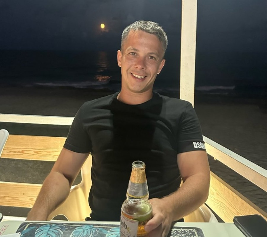 Man sitting outdoors at night holding a beer, ocean in background.