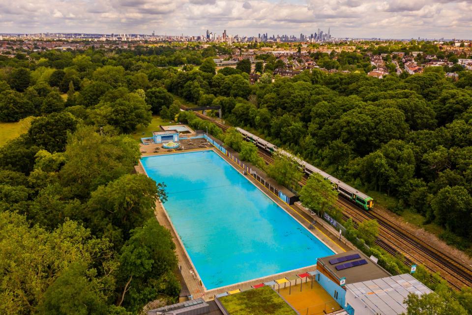 Aerial view of a closed swimming pool next to a train track, with a city skyline in the distance.