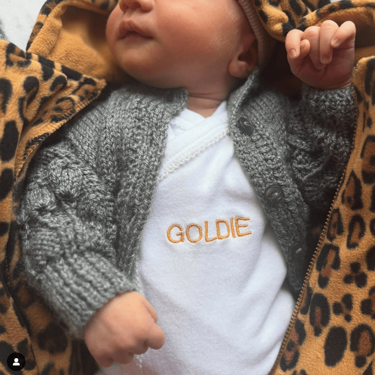 Baby wearing a gray cardigan and white onesie that says "Goldie".