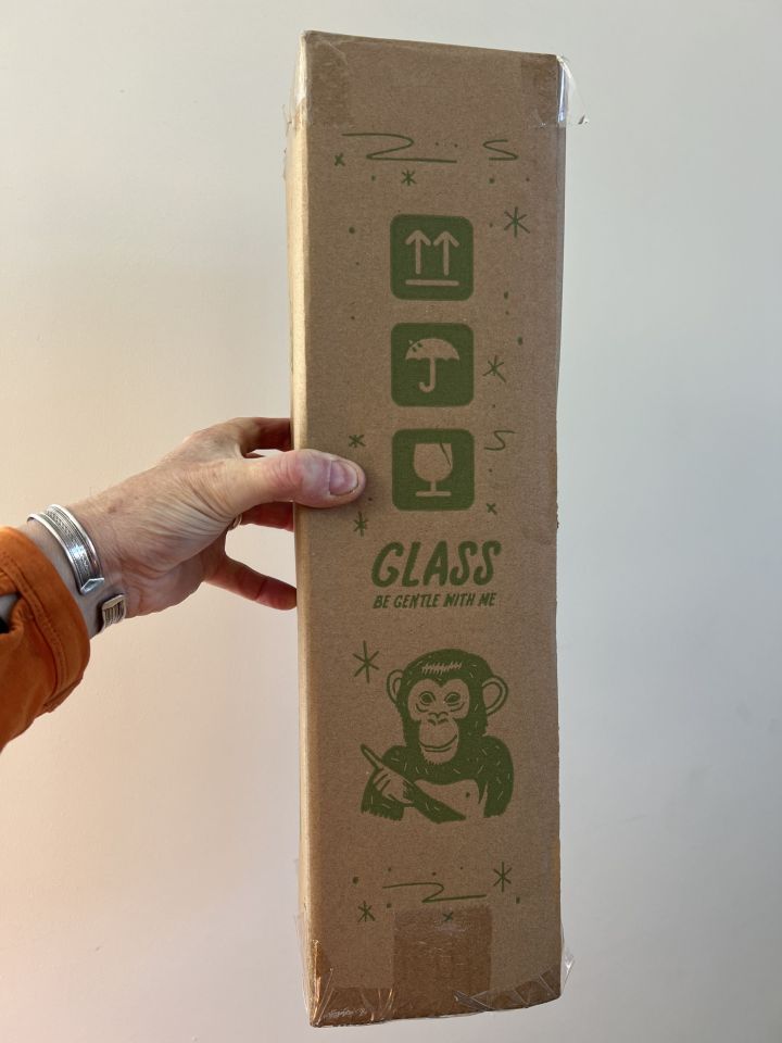 A hand holding a cardboard box labeled "Glass, be gentle with me," featuring a chimpanzee illustration and fragile item symbols.