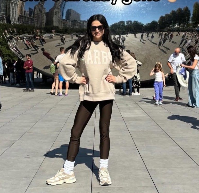 Woman standing in front of a large sculpture, wearing a beige sweatshirt and black leggings.