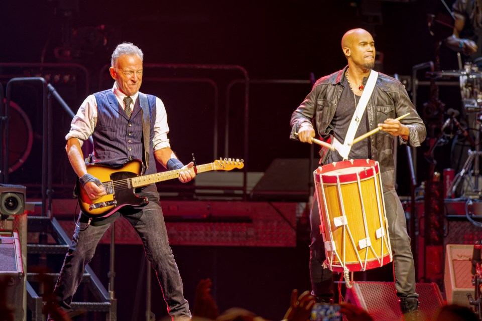 Photo of Bruce Springsteen playing guitar and a percussionist playing a drum during a concert.