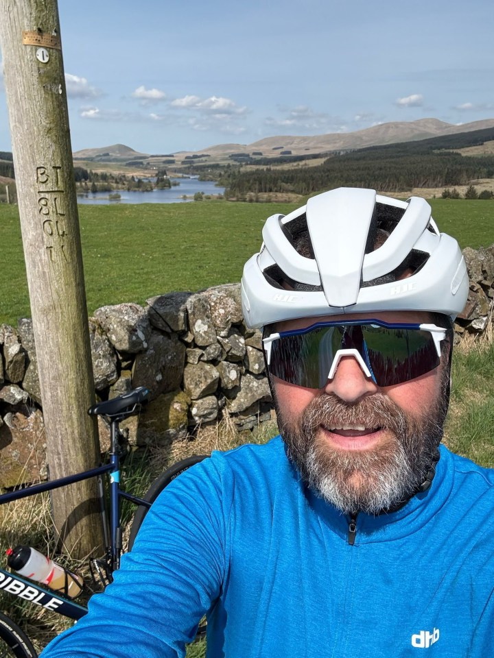 Man in cycling gear taking a selfie with his bike and a scenic view in the background.