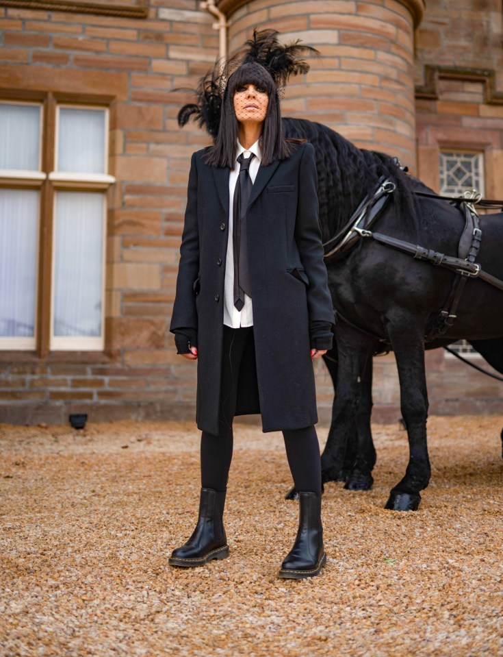 Claudia Winkleman standing in front of a black horse and carriage.