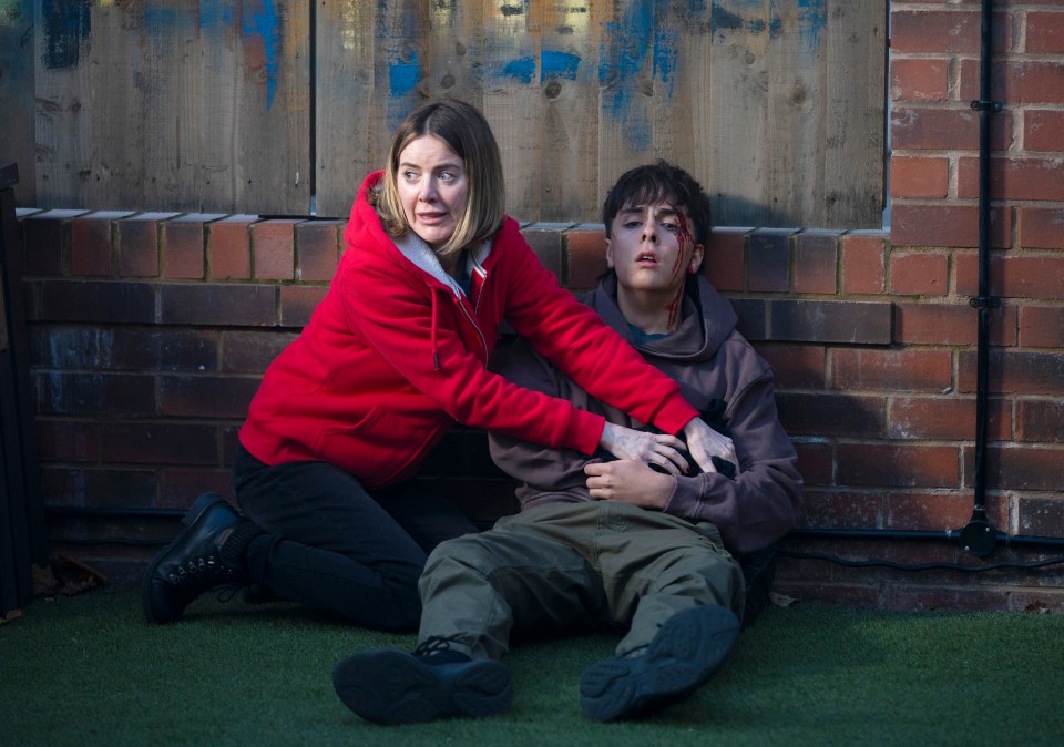 A woman cradles a bleeding teenage boy against a brick wall.