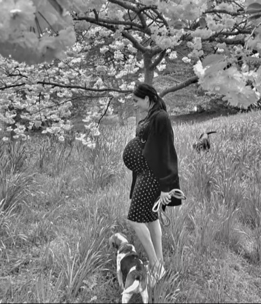Black and white photo of a pregnant woman walking a dog under a blossoming tree.