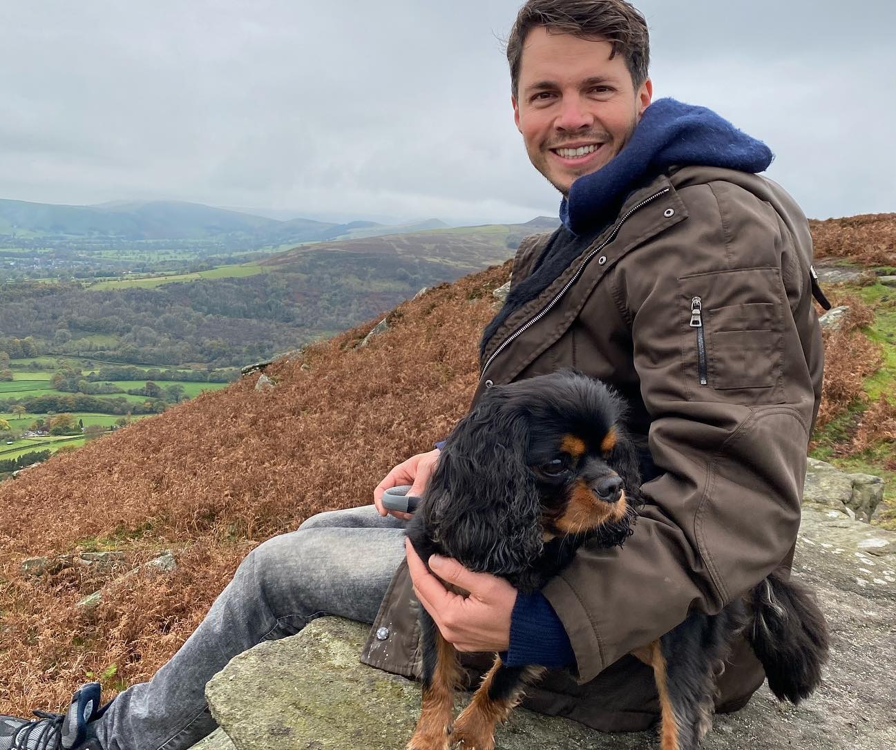 Man holding a Cavalier King Charles Spaniel dog while sitting on a rock overlooking a valley.
