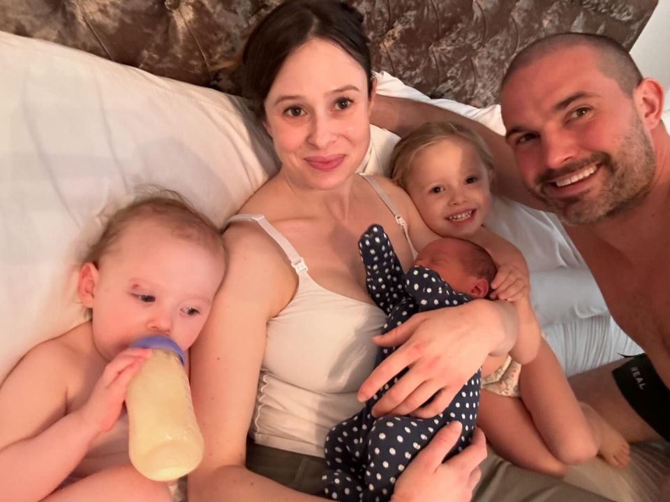 Family of five in bed; parents hold newborn and toddler, while older daughter sits between them.