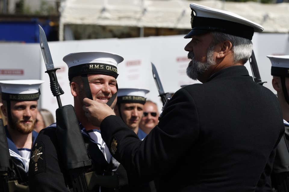 A man in a navy uniform adjusting the neckerchief of a sailor during a ceremony.